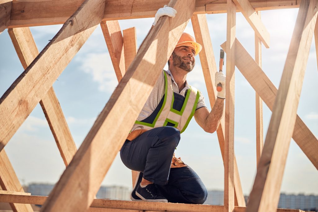 Construction worker in hard hat and safety vest kneeling on wooden roof trusses at a framing job site